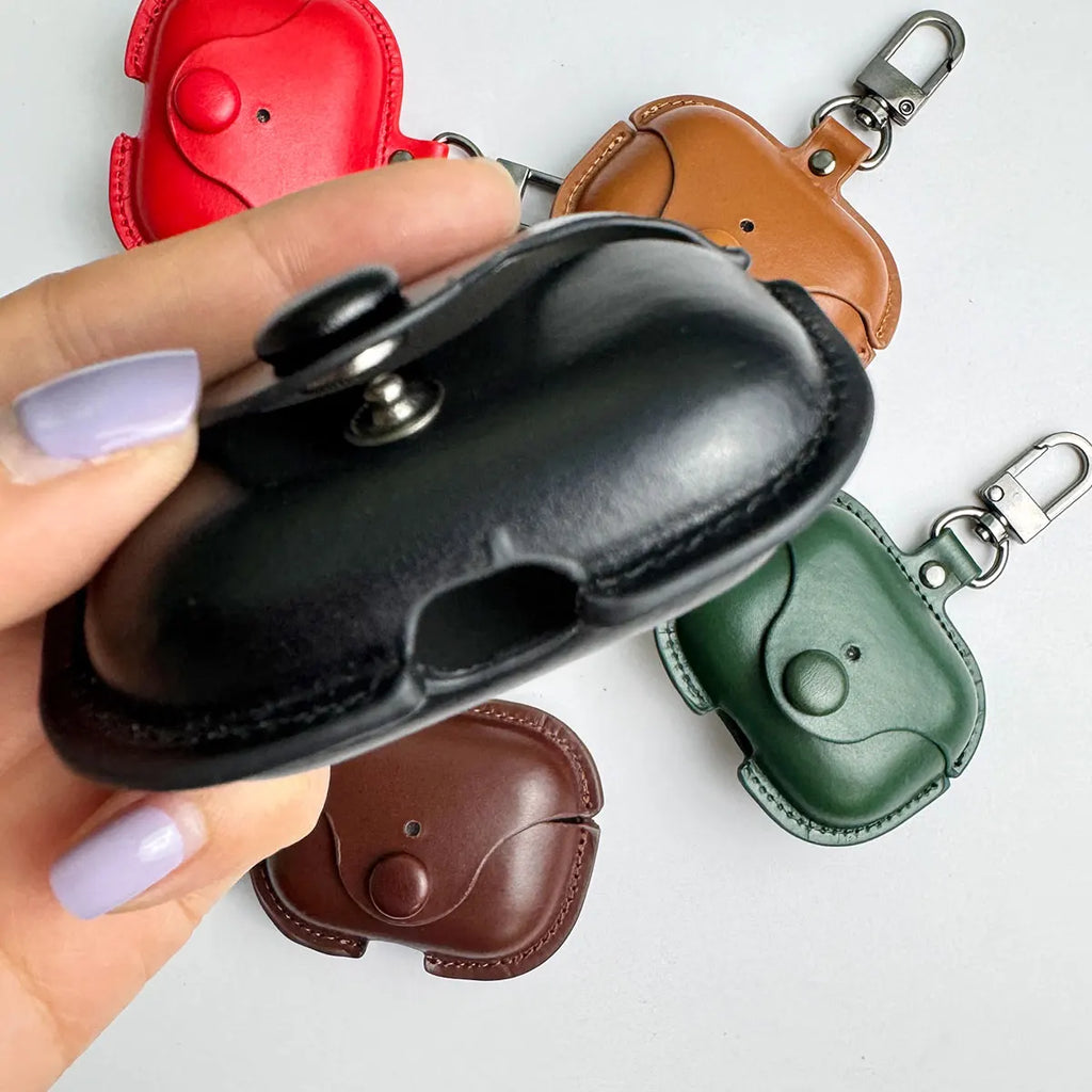 Hand holding a black leather AirPods case with popper fastening and keychain; other colours available on white surface.