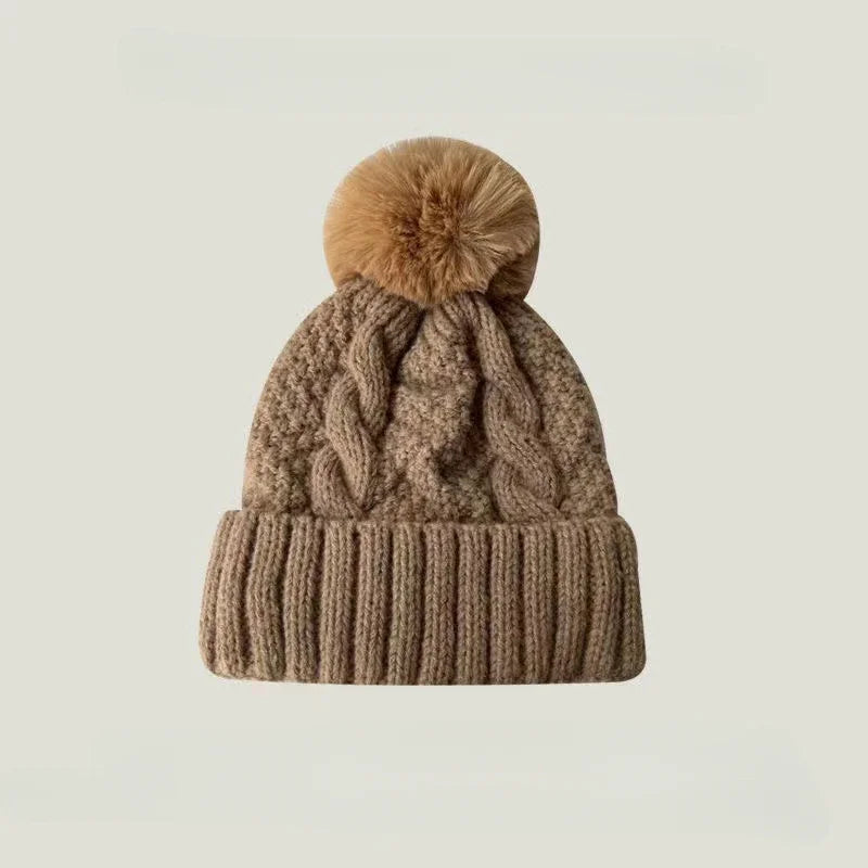 Beige cable-knit beanie hat with a ribbed cuff and a matching fluffy faux fur pom-pom on top.