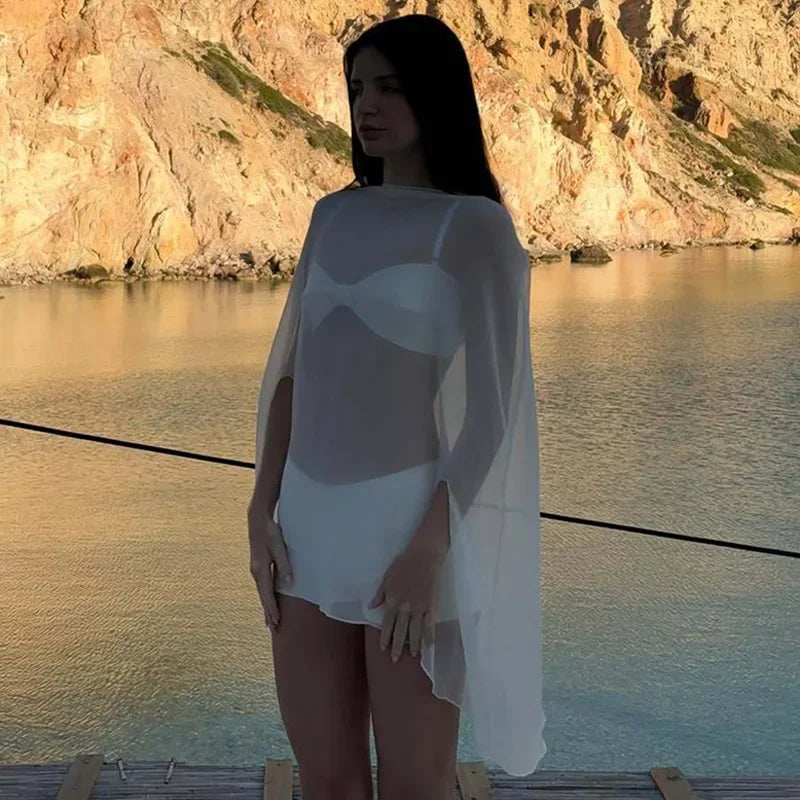 Woman wearing a sheer white chiffon poncho cover-up over a matching bikini set against a coastal backdrop.