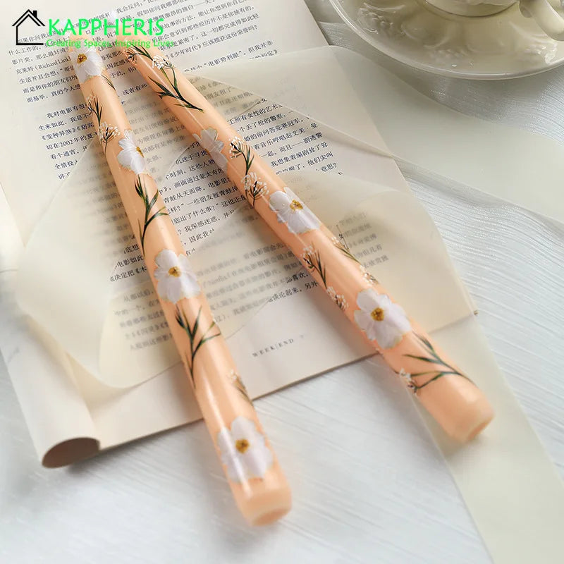 Two tapered peach candles decorated with delicate white floral and green leaf illustrations, resting on a paper book.