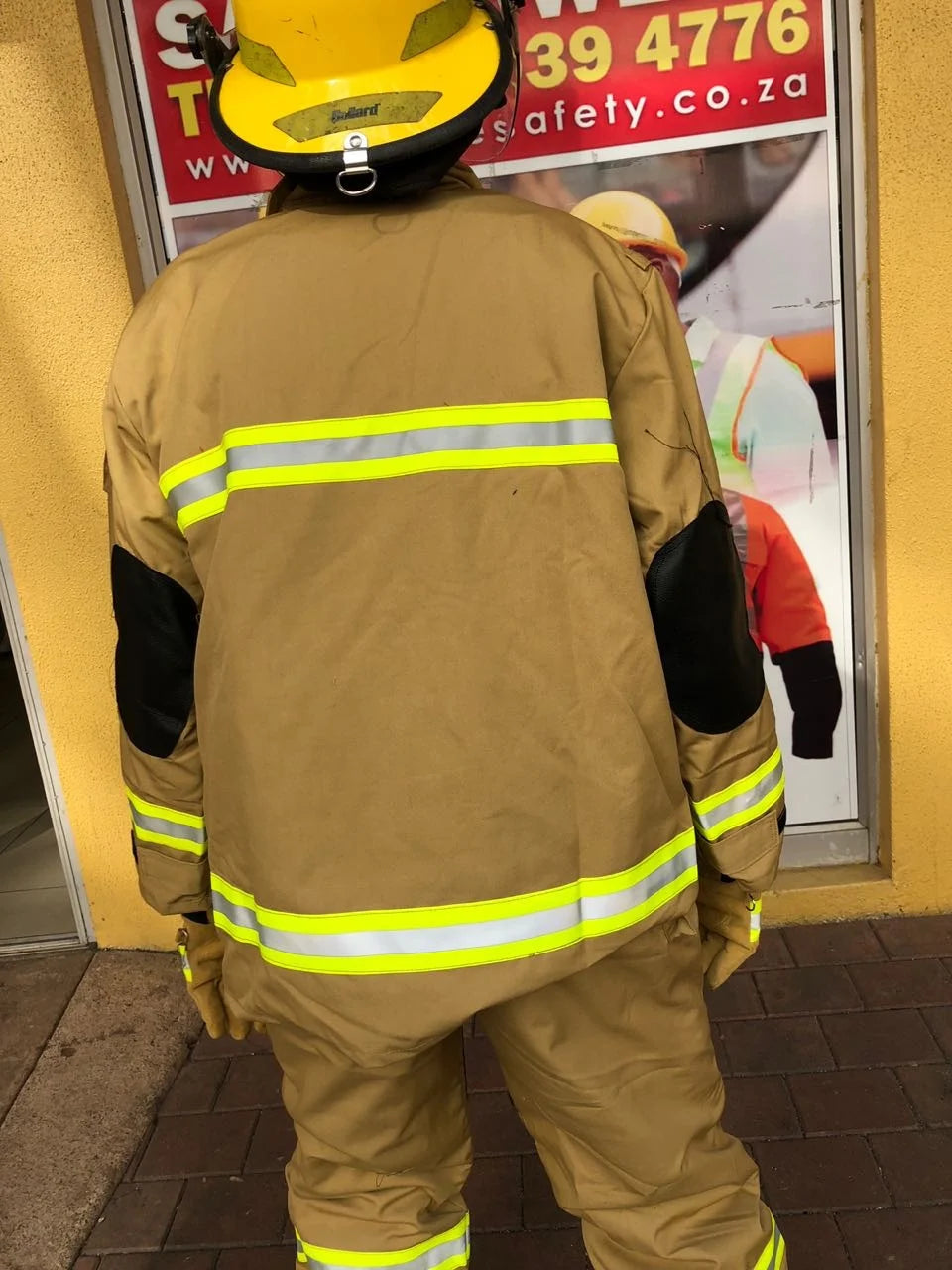 Tan canvas firefighter uniform with black elbow patches, yellow reflective strips, and a yellow safety helmet.
