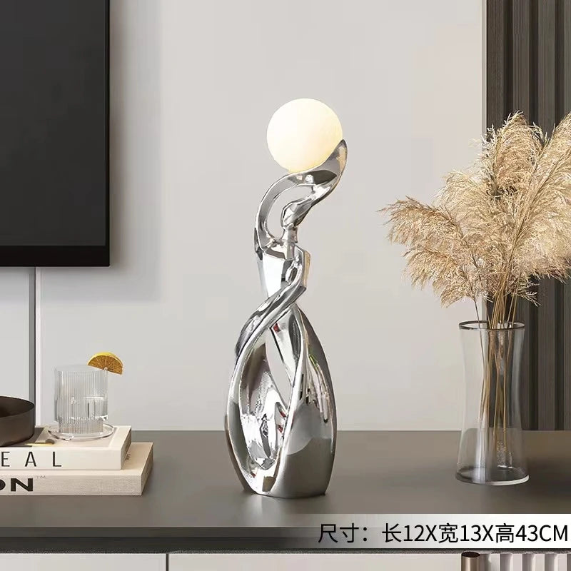 Abstract silver-finish resin ornament featuring a swirling sculptural base topped with a glowing white spherical light.