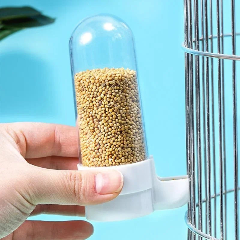 Hand holding a clear plastic, cylindrical bird feeder filled with seeds, attached to a metal birdcage.