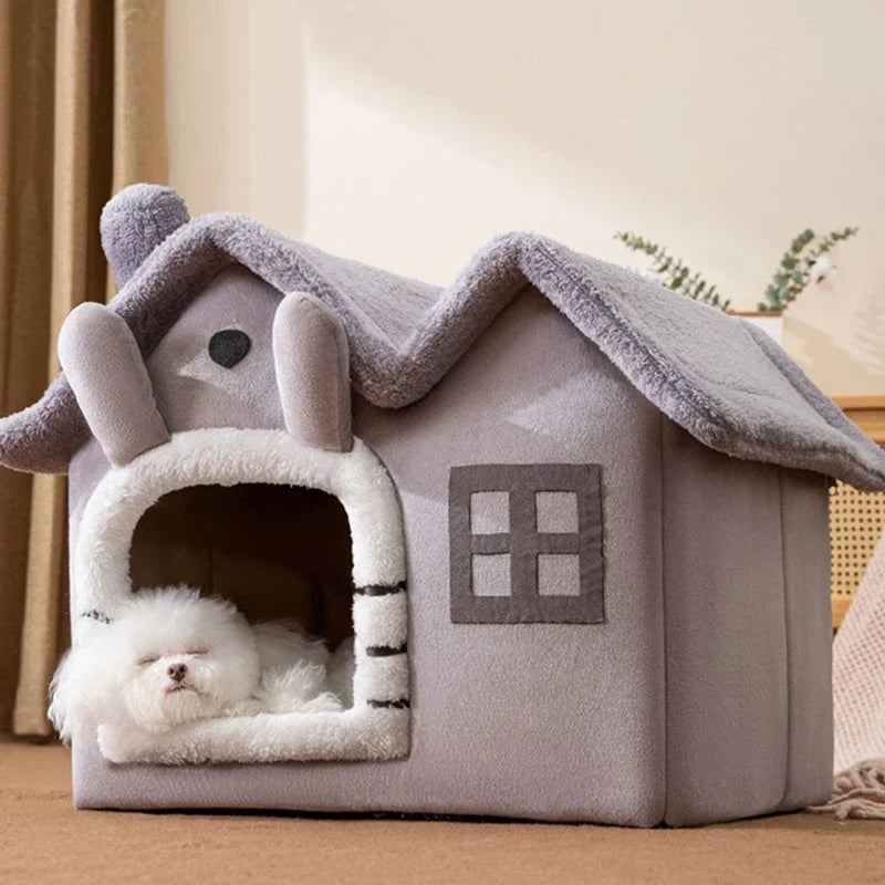 Gray house-shaped pet bed with a fluffy roof and chimney, featuring a window and a small white dog sleeping inside.