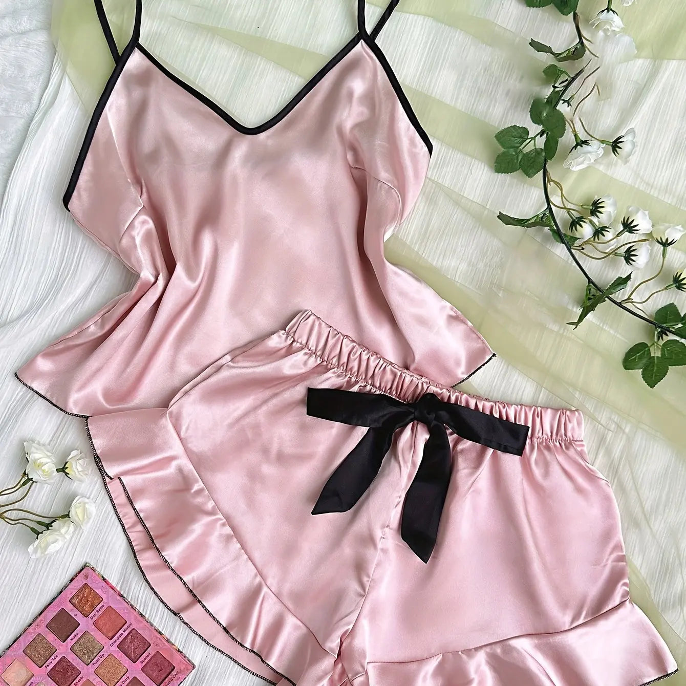 Pink satin camisole pyjama set with black trim, ruffled shorts, and a waist bow on a white background.
