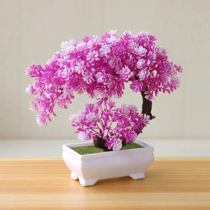 An artificial pink bonsai tree in a rectangular white pot, set on a wooden table against a plain light background.