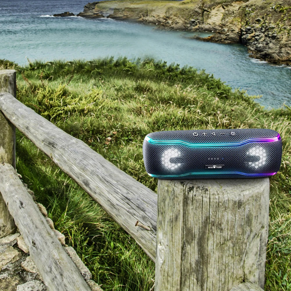 Portable black Bluetooth speaker with vibrant LED lights, resting on a wooden fence post overlooking the sea.