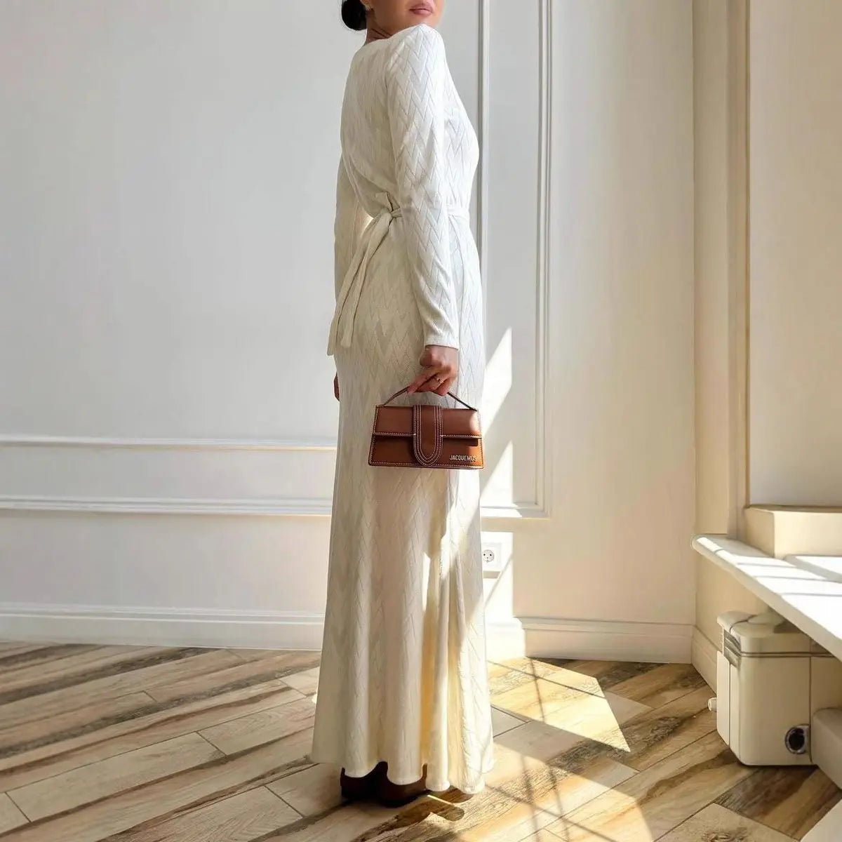 Elegant cream maxi dress with long sleeves, tie-waist detail, and subtle chevron texture, worn with a brown handbag.