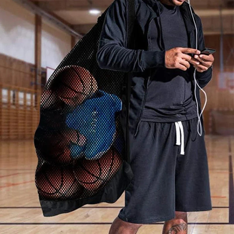 A man in a gym holding a black mesh sports bag filled with basketballs and a blue item.