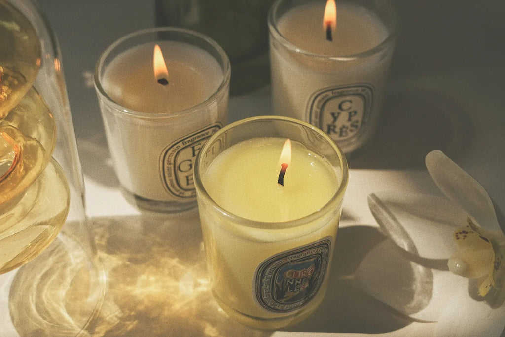 Three lit scented candles in clear glass tumblers with elegant labels, set against a warm, soft-focus background.