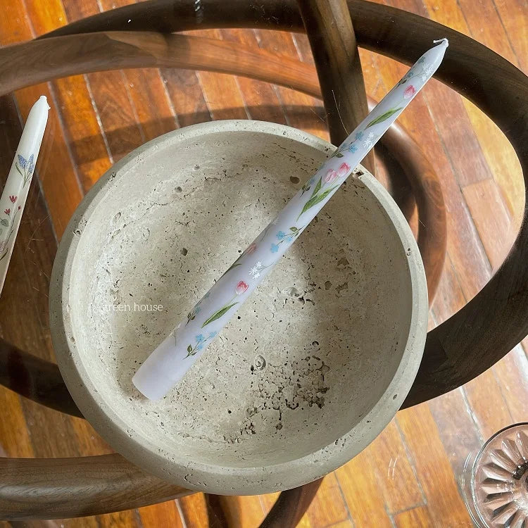 Taper candle with a pale grey finish and delicate floral print, resting in a textured stone bowl.