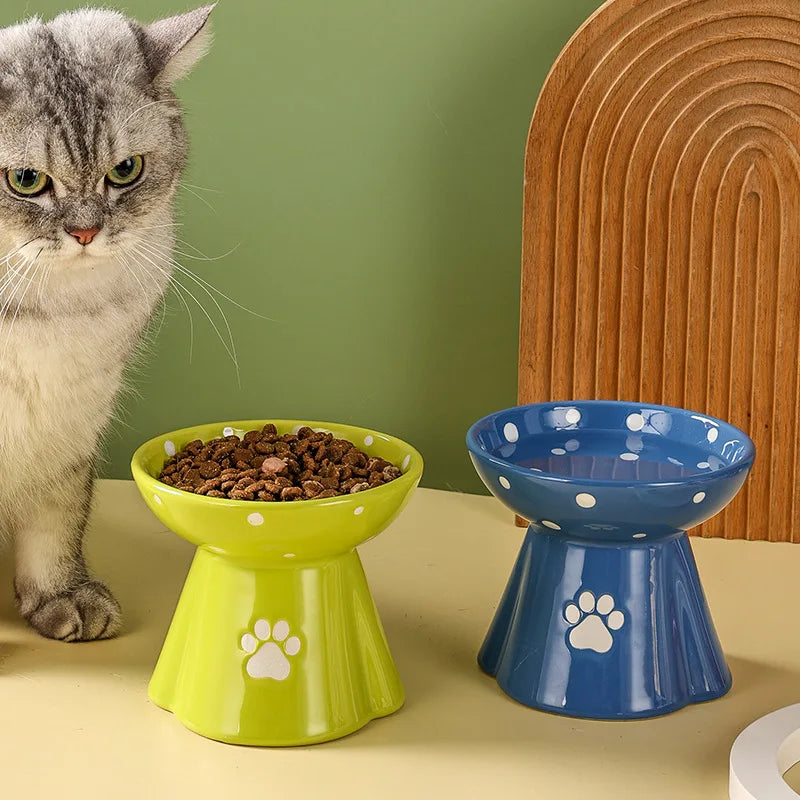 Green and blue elevated ceramic cat bowls with white polka dots and paw print designs, set on a cream surface.