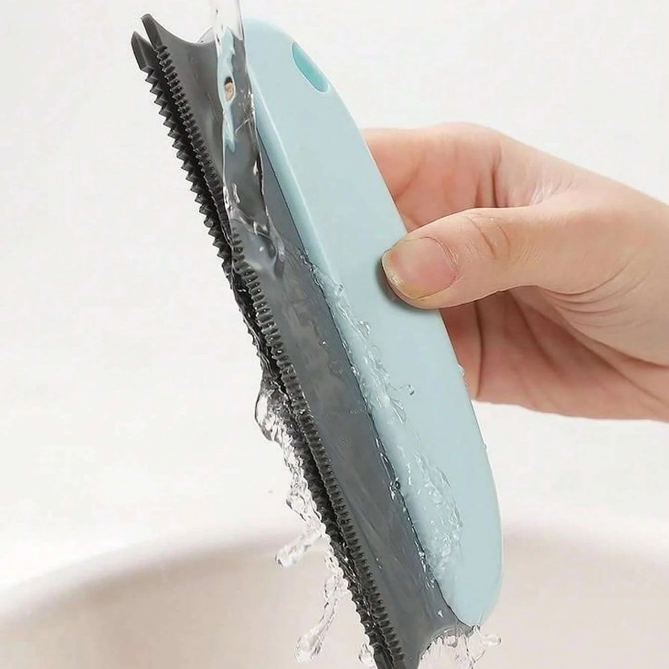 Handheld blue and grey cleaning tool with a ribbed silicone edge, being rinsed under running water.