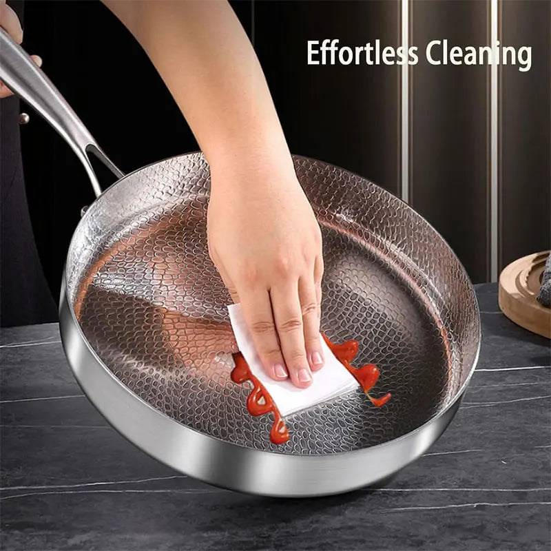 A hand wipes red sauce from a textured, stainless steel frying pan with a paper towel.