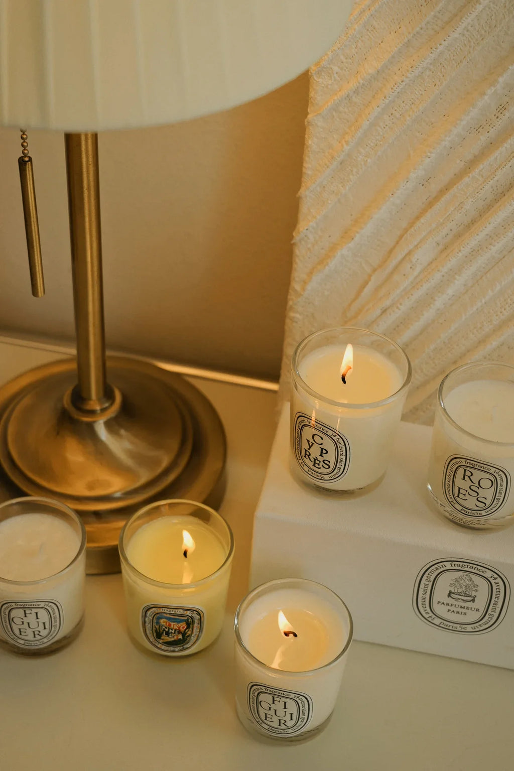 Several luxury scented candles in glass jars, some lit, arranged on a white stand next to a gold lamp.