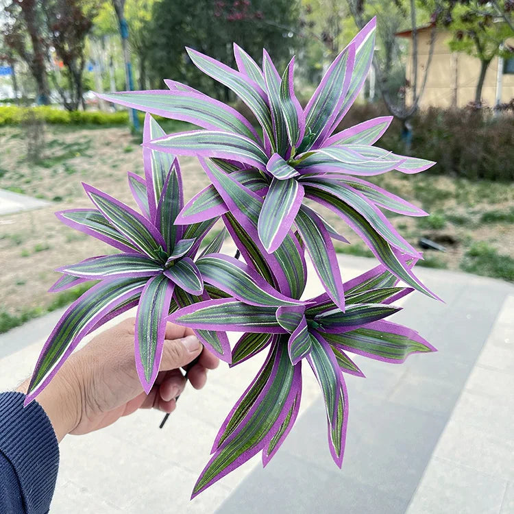 Hand holding a bunch of artificial plants with long, pointed leaves featuring purple edges and striped green centers.