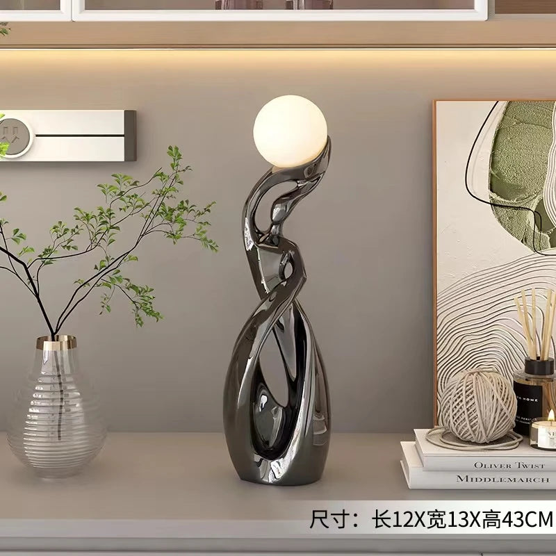 Abstract dark chrome resin floor ornament featuring a smooth, twisting silhouette topped with a glowing spherical light.