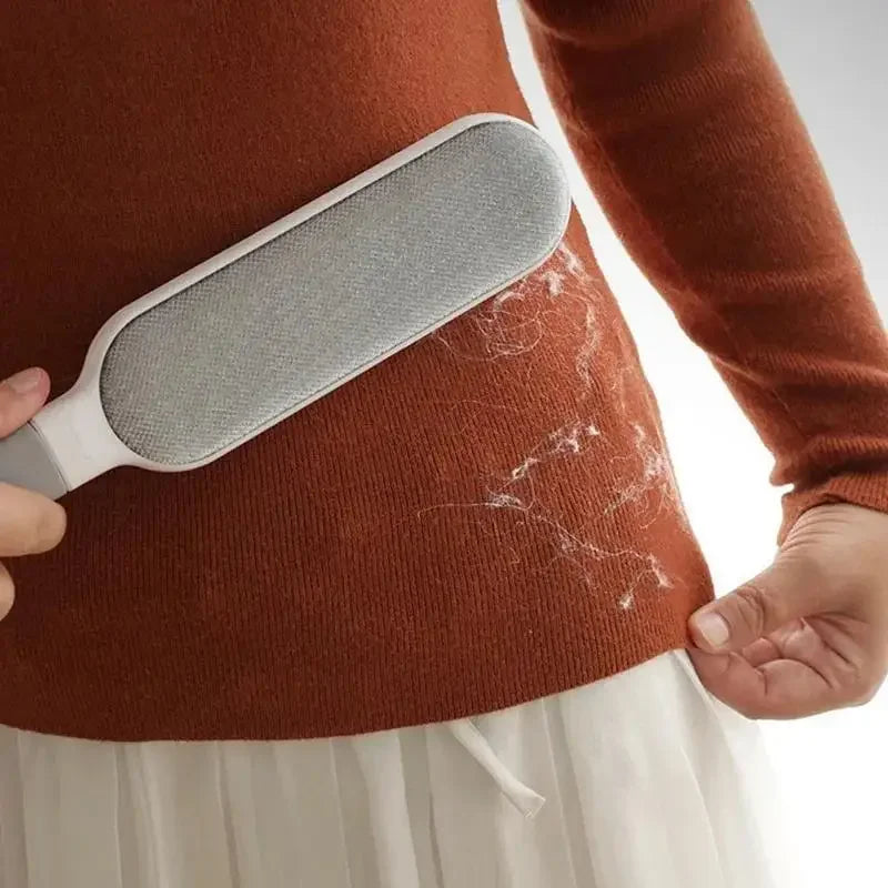 A grey, handheld fabric lint remover being used on a rust-orange knitted jumper covered in white lint and hair.