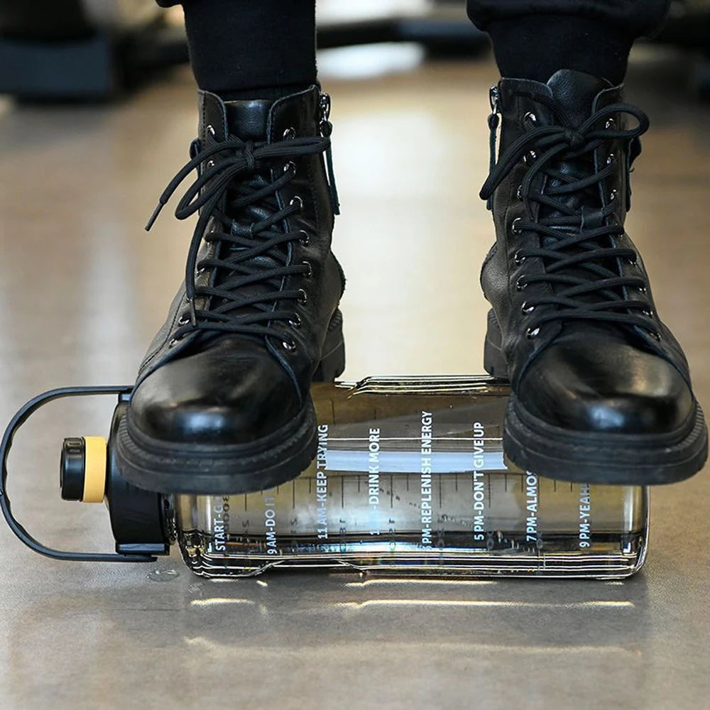 Black boots standing on a large, clear motivational water bottle with time markers and a yellow cap detail.
