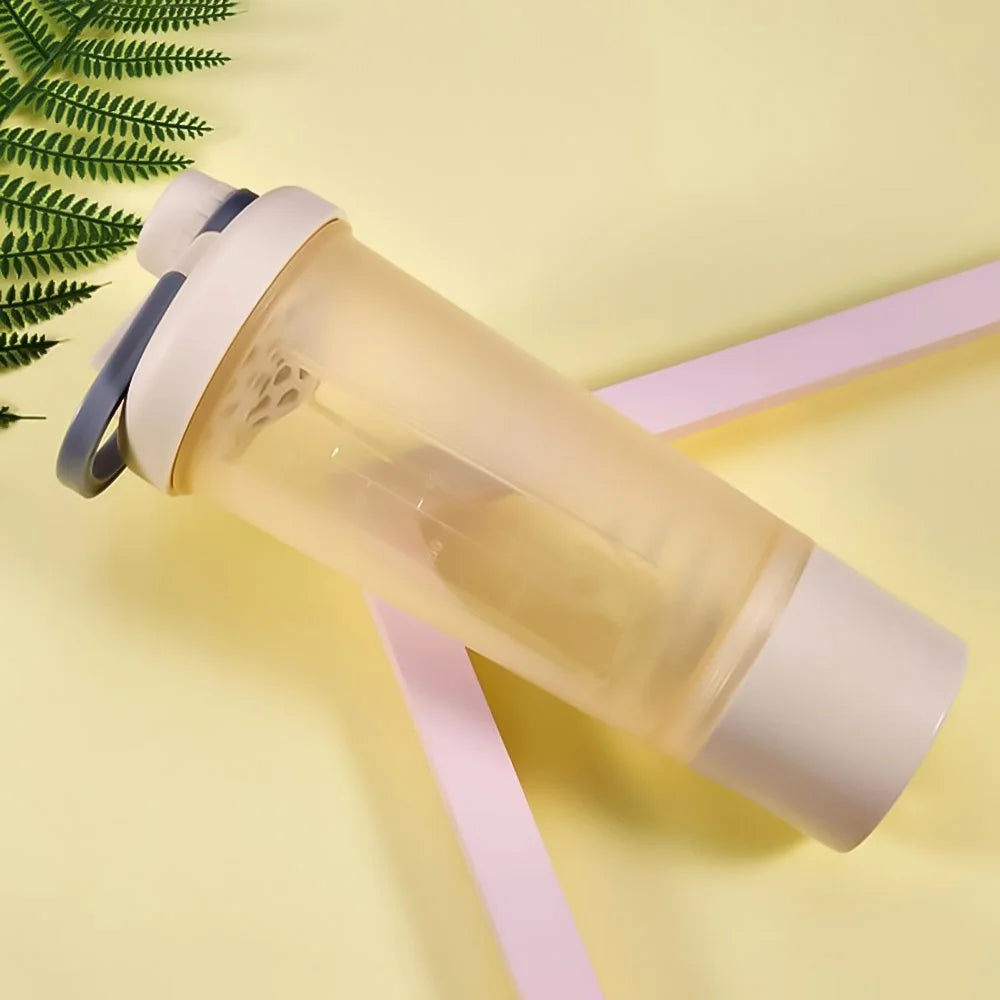 A translucent shaker bottle with a cream-colored lid, grey handle, and base on a yellow background with fern foliage.