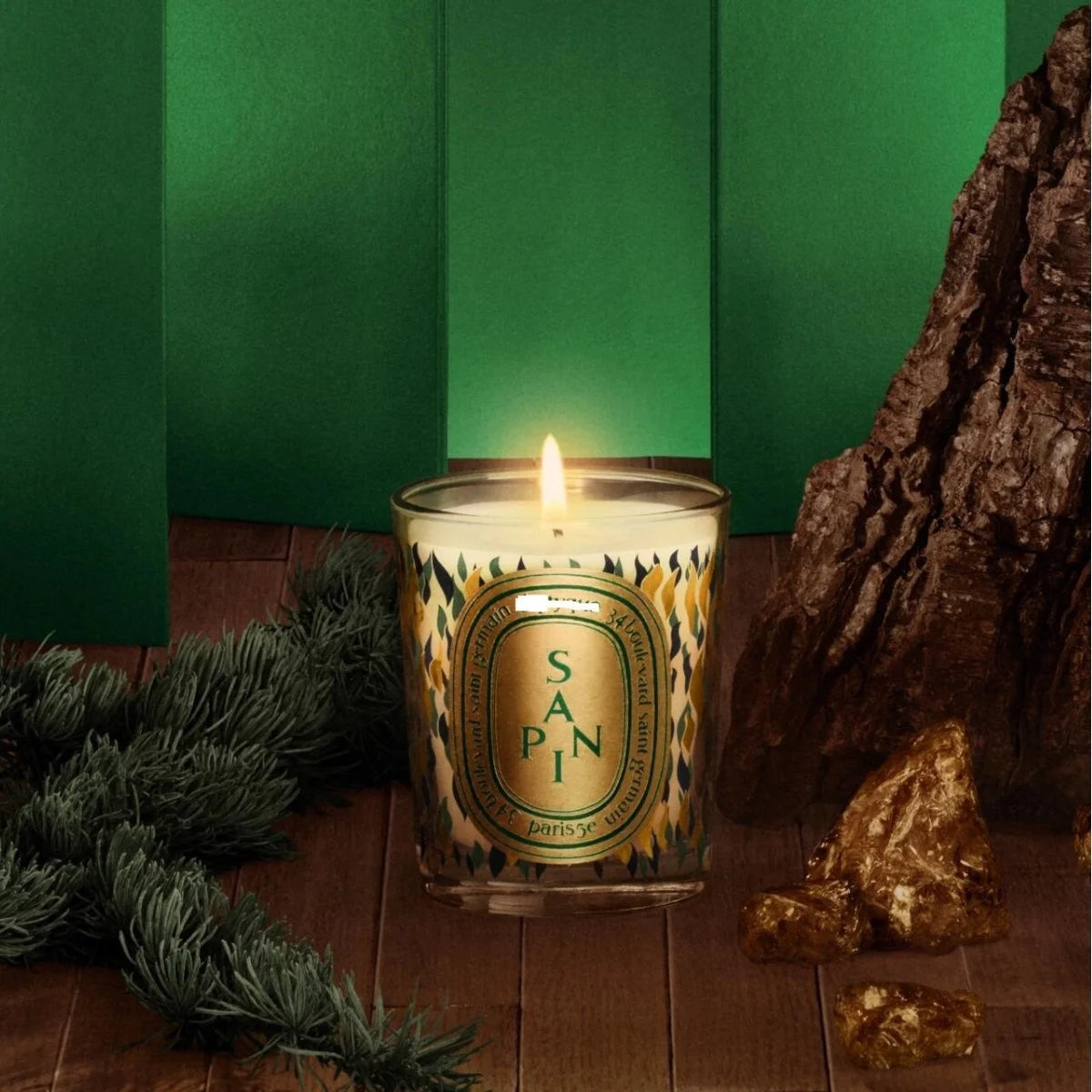 Lit scented candle in a glass jar with decorative pine patterns, set against a green backdrop with fir needles and resin.