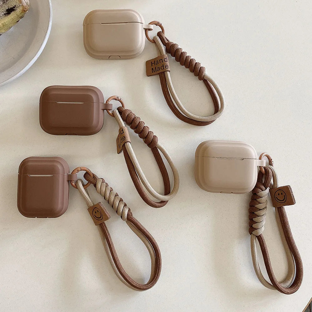 Matte silicone AirPods cases in neutral brown tones, featuring decorative braided wrist straps with charm details.