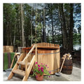 A wooden hot tub with stairs and a smoking chimney stands outdoors surrounded by trees, near a lake.