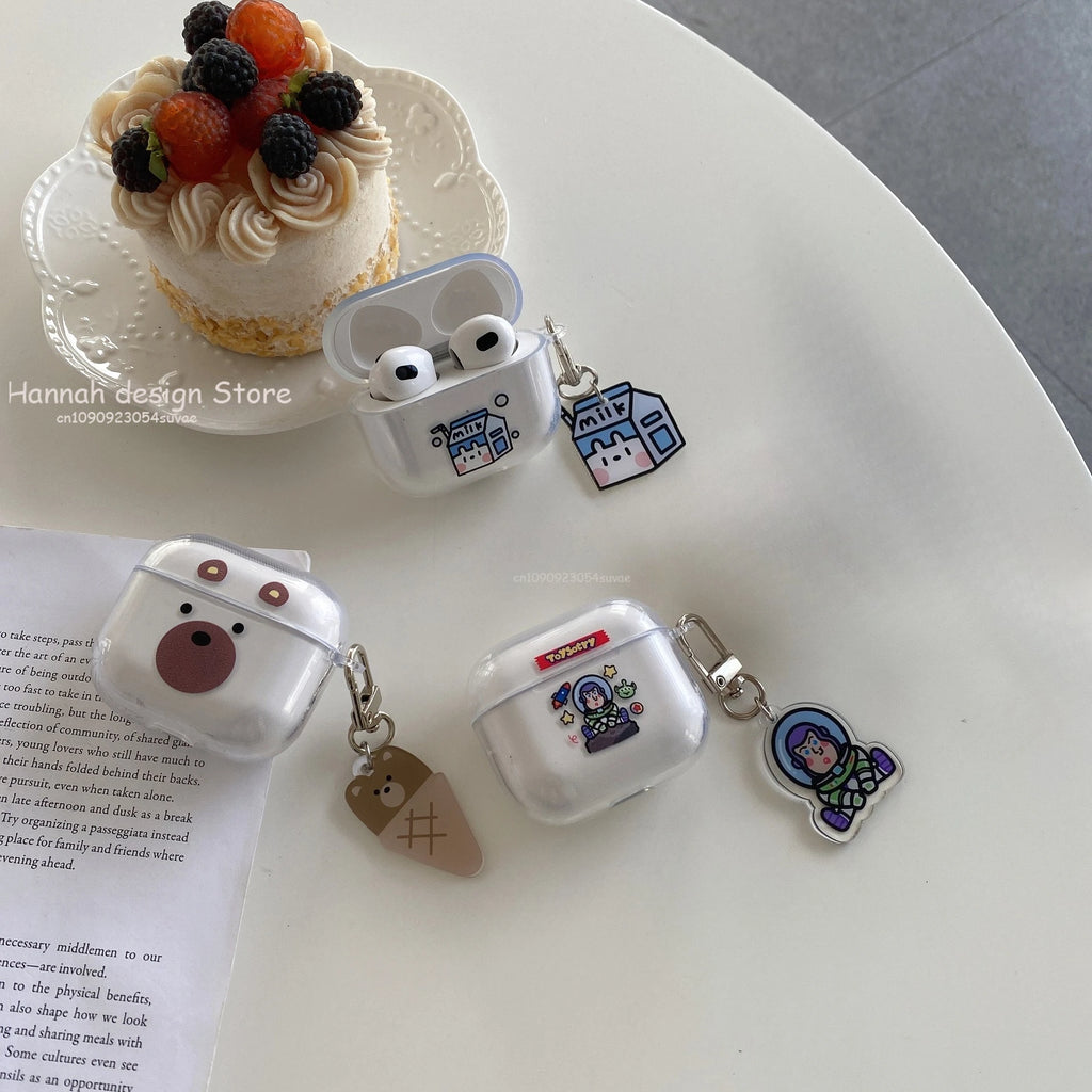 Clear AirPods 3rd Gen cases with cute character prints and matching hanging charms, on a table with a mini cake.
