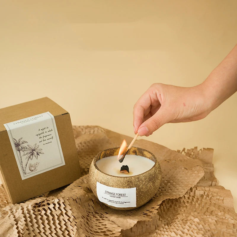 A hand lights a soy wax candle in a rustic, natural coconut shell bowl beside a kraft paper gift box.