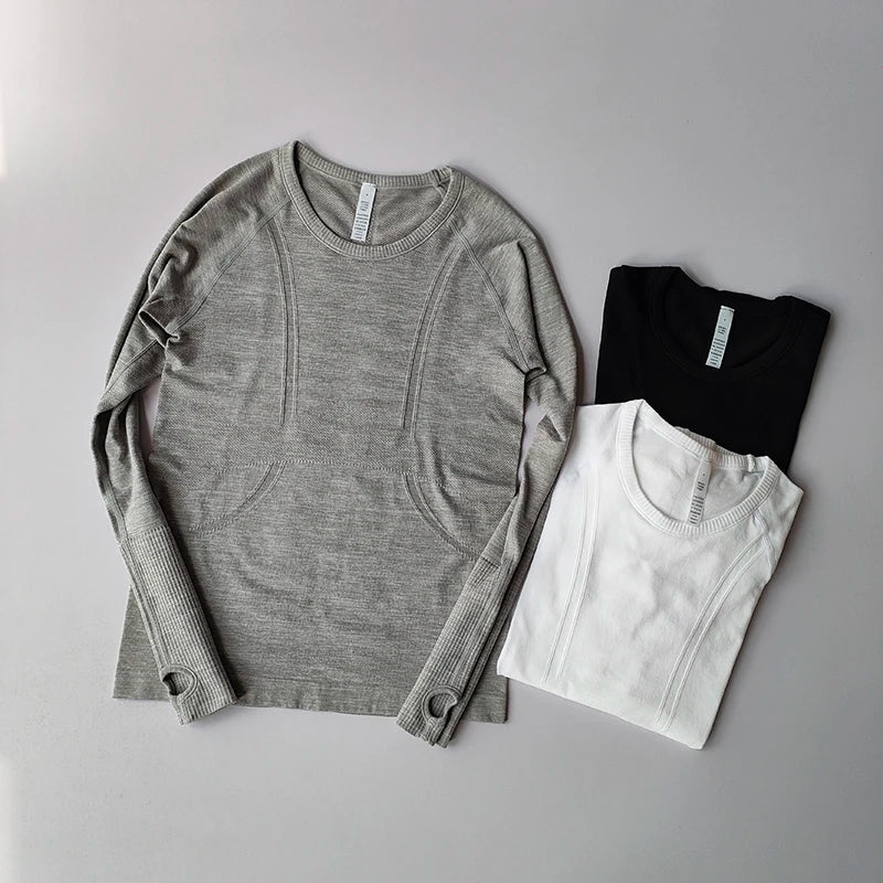 Three seamless long-sleeve sports tops in grey, black, and white with ribbed cuffs and thumb holes.