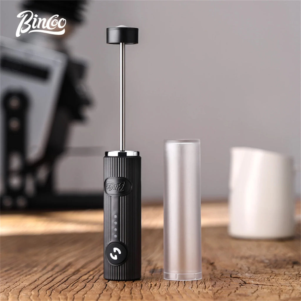 Handheld electric milk frother with a black ridged handle and a metal whisk, standing next to a clear protective cover.