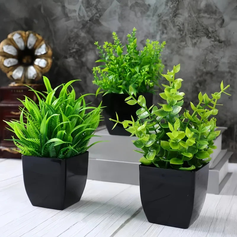Three small, artificial green plants in square black pots arranged on a white wooden surface against a grey backdrop.