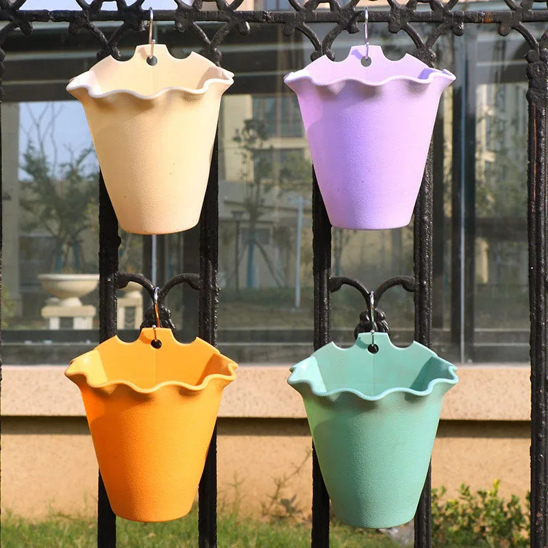 Four pastel-colored, scalloped-edge plastic planters hang from metal hooks on a black iron fence.