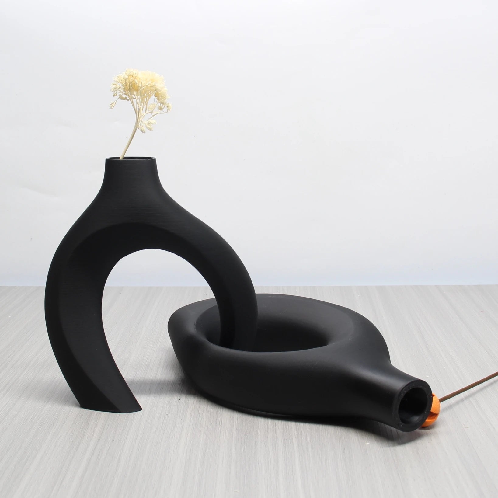 Two matte black, sculptural ceramic vases interlinked in a modern bohemian design with a dried flower stem.