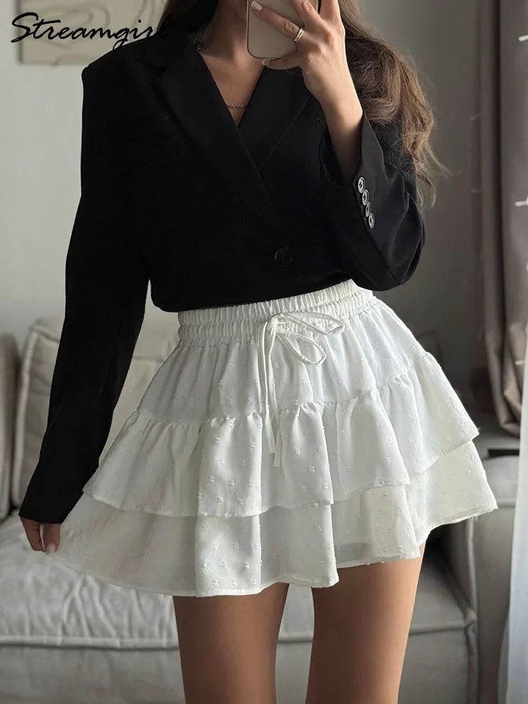 White A-line mini skirt with a smocked drawstring waist, layered ruffles, and Swiss dot texture.