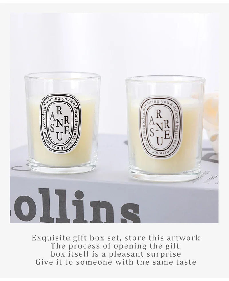 Two cream-coloured scented candles in clear glass tumblers with oval labels, displayed on a grey book.