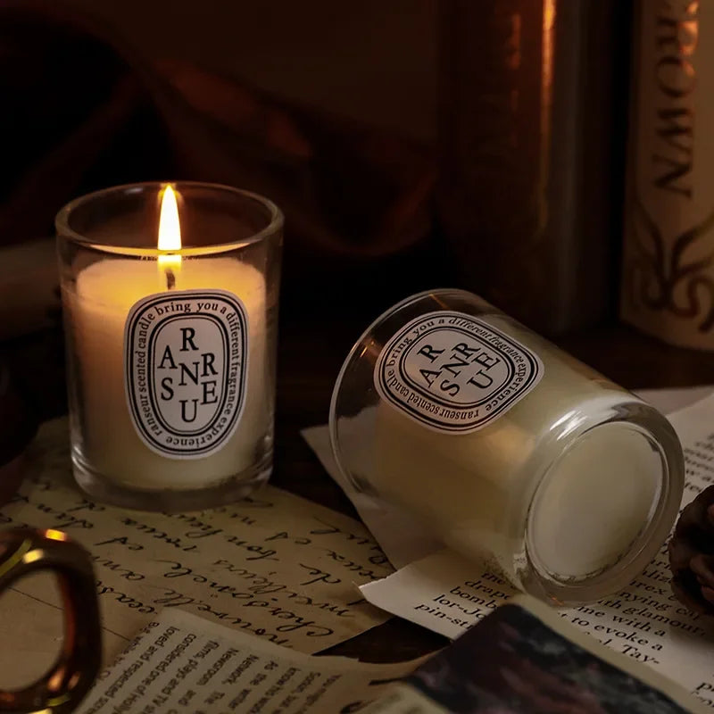 Two white scented candles in clear glass tumblers with oval labels on a vintage, literary-themed tabletop.