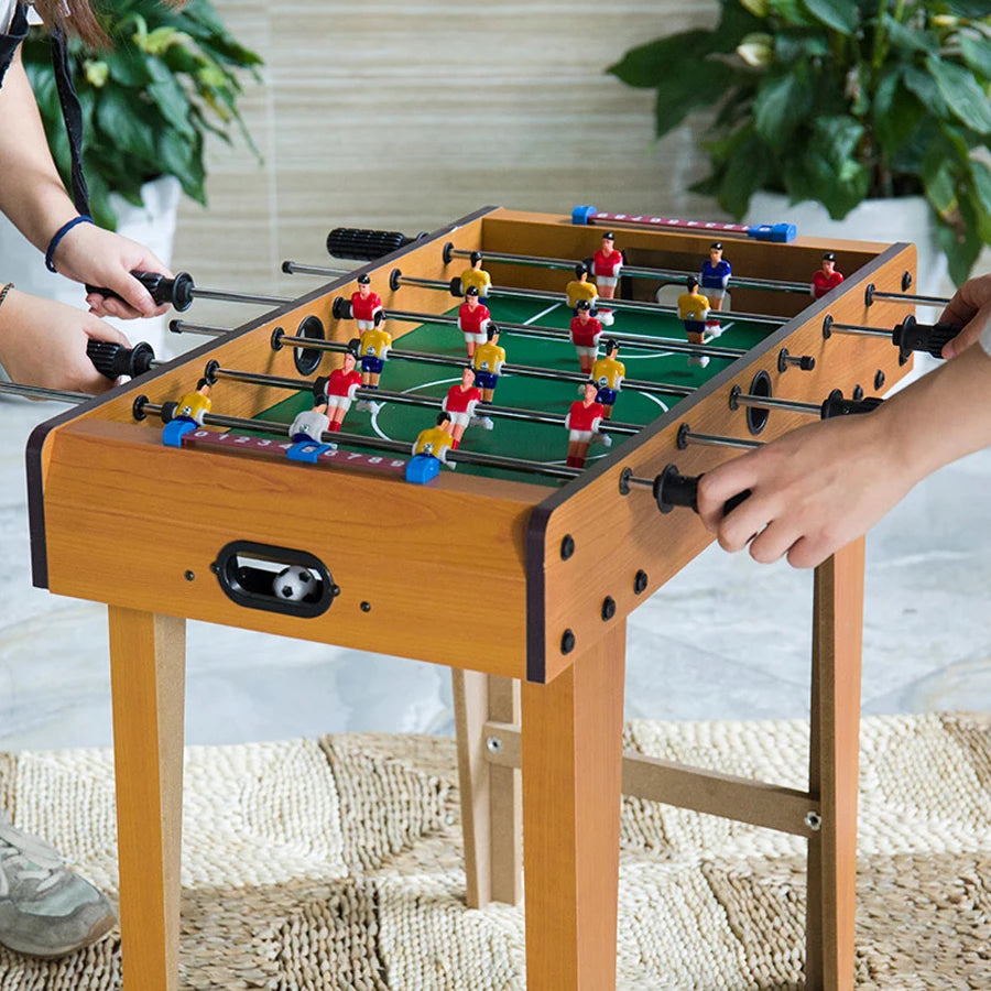 Wooden foosball table with green pitch, sliding score bars, and players in red, yellow, and blue kits.