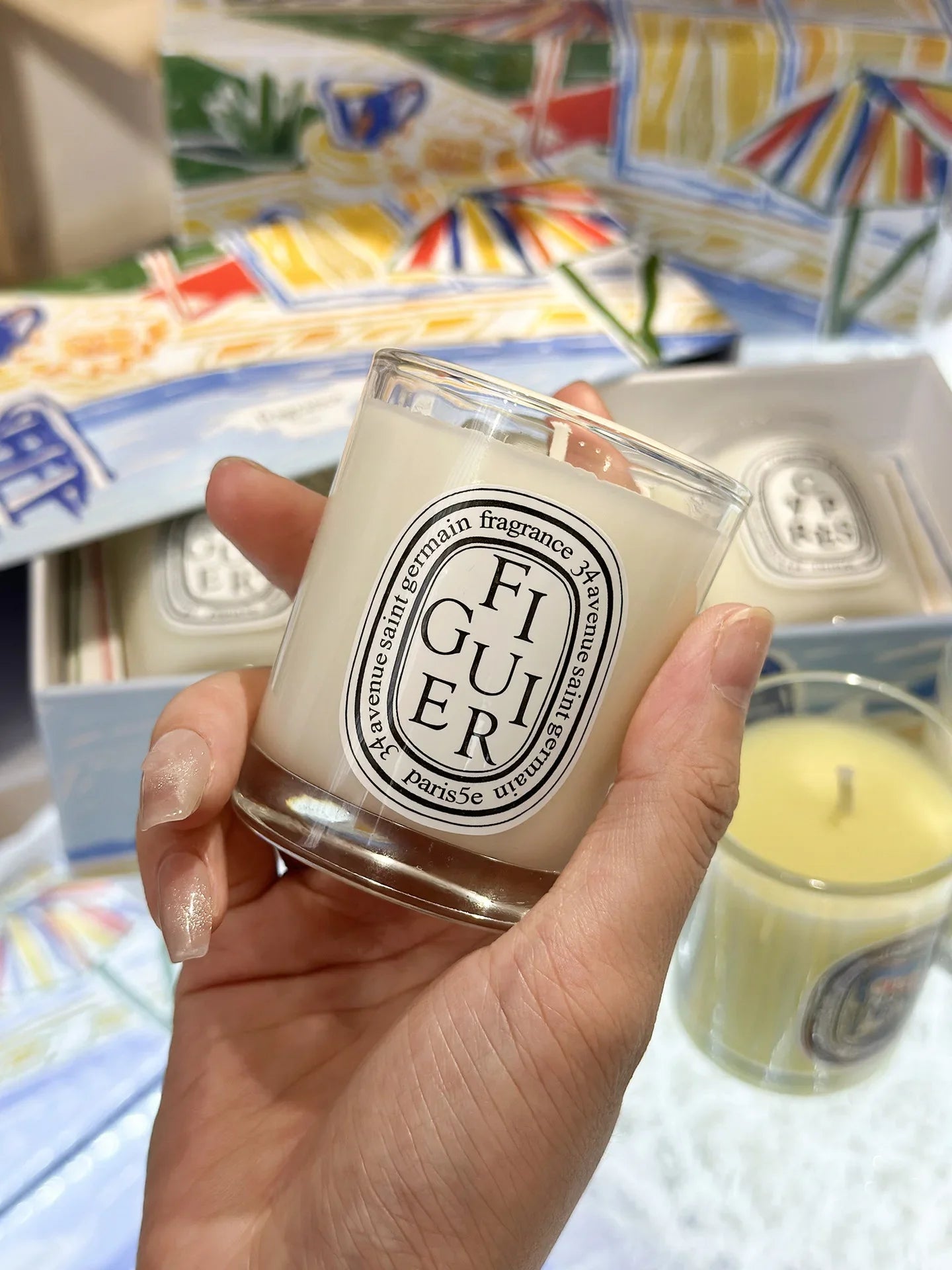 Hand holding a white scented candle in a clear glass jar with an oval white label, presented in an illustrated gift box.