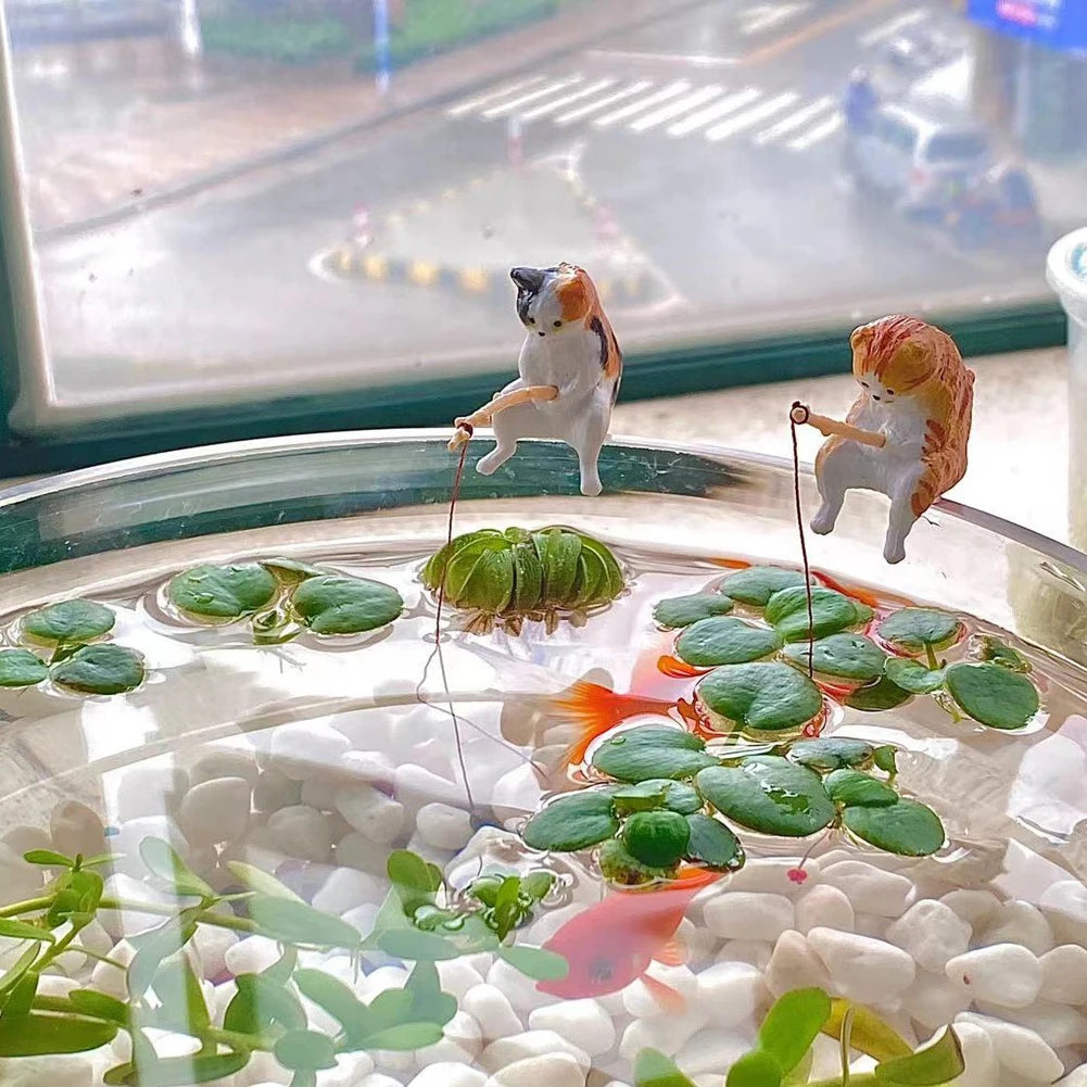 Two miniature cat ornaments, one ginger and one calico, perched on an aquarium rim, holding small fishing rods.