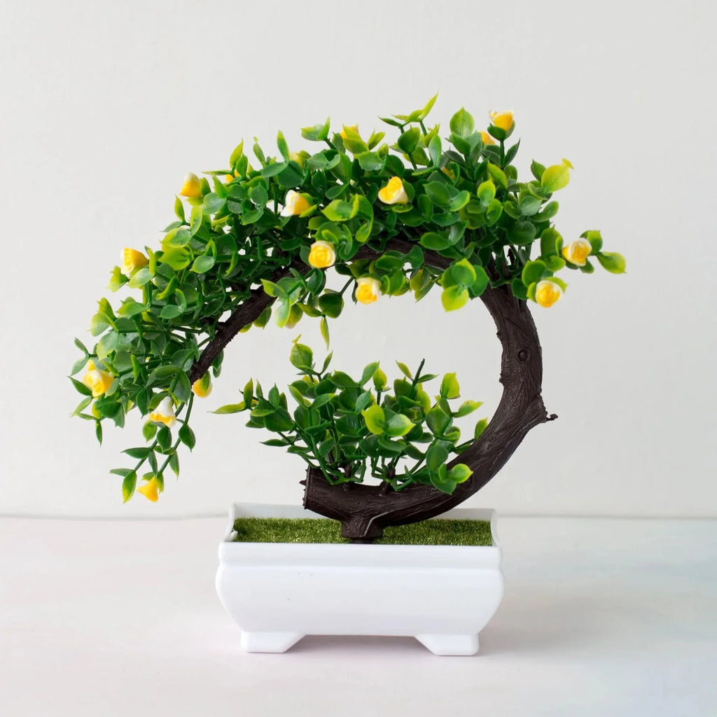 Artificial bonsai tree with a curved brown trunk, green leaves, and tiny yellow flowers in a rectangular white pot.