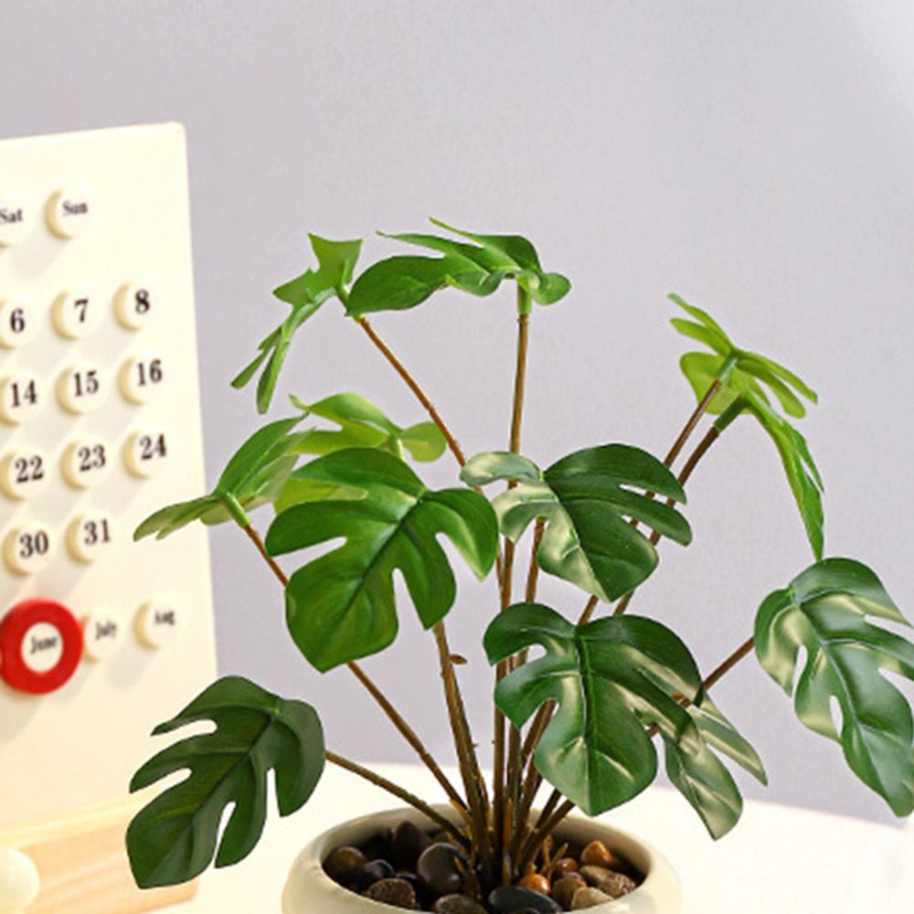 Artificial monstera plant with deep green split leaves in a white ceramic pot filled with small decorative stones.