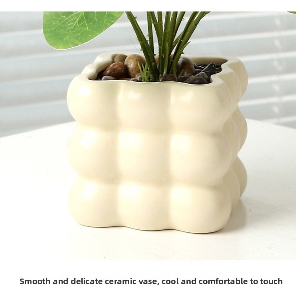 Artificial plant in a cream-colored, square, bubble-textured ceramic pot topped with decorative pebbles.